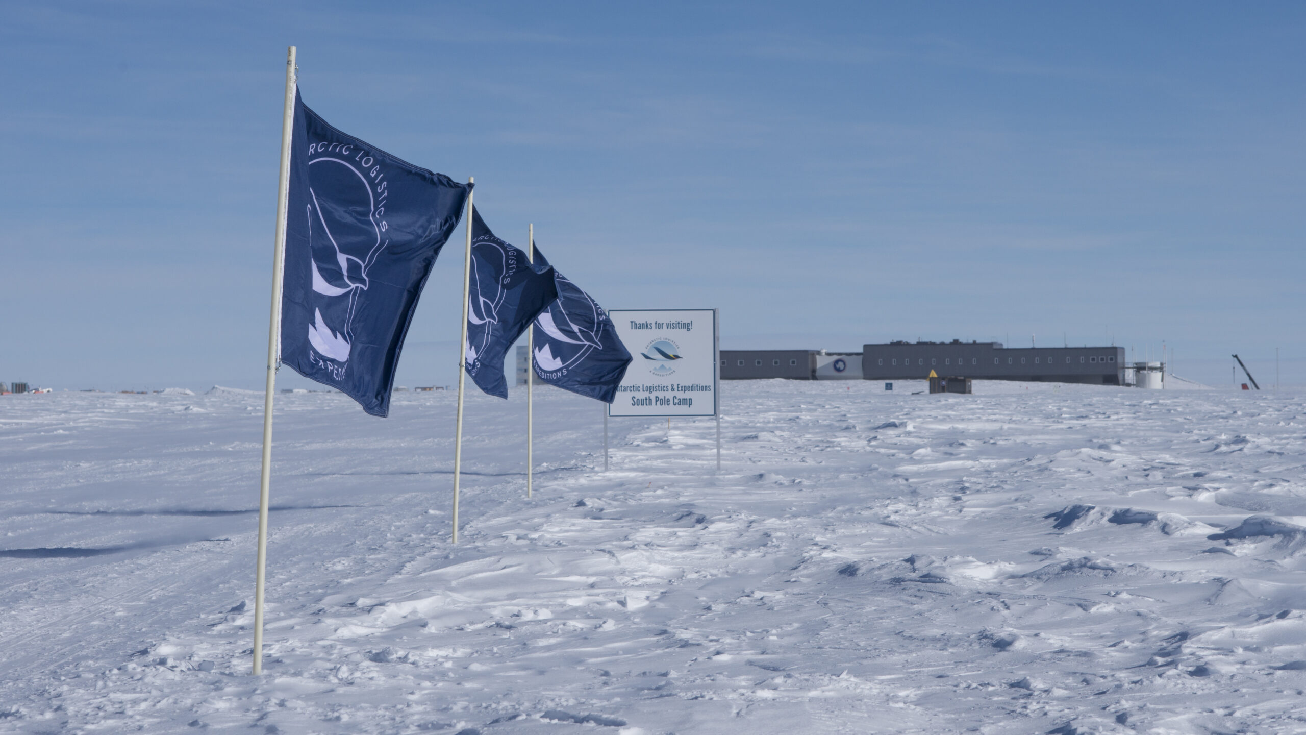 ALE flags and a sign, mark the edge of ALE's South Pole Camp. Amundsen-Scott South Pole station in the background.